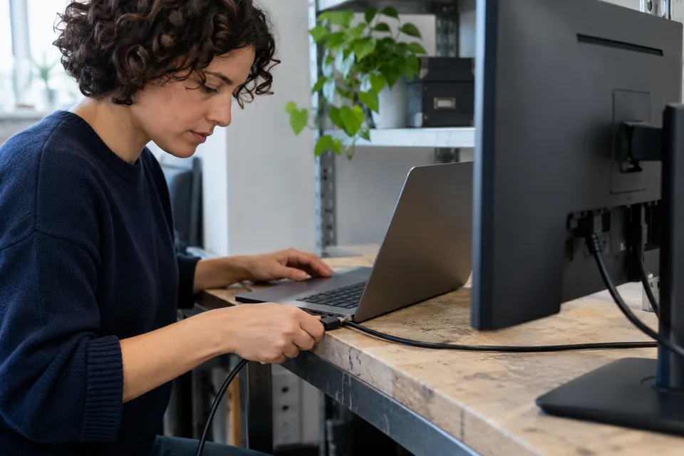 Persoon sluit een kabel aan op een laptop bij een eenvoudige werktafel.